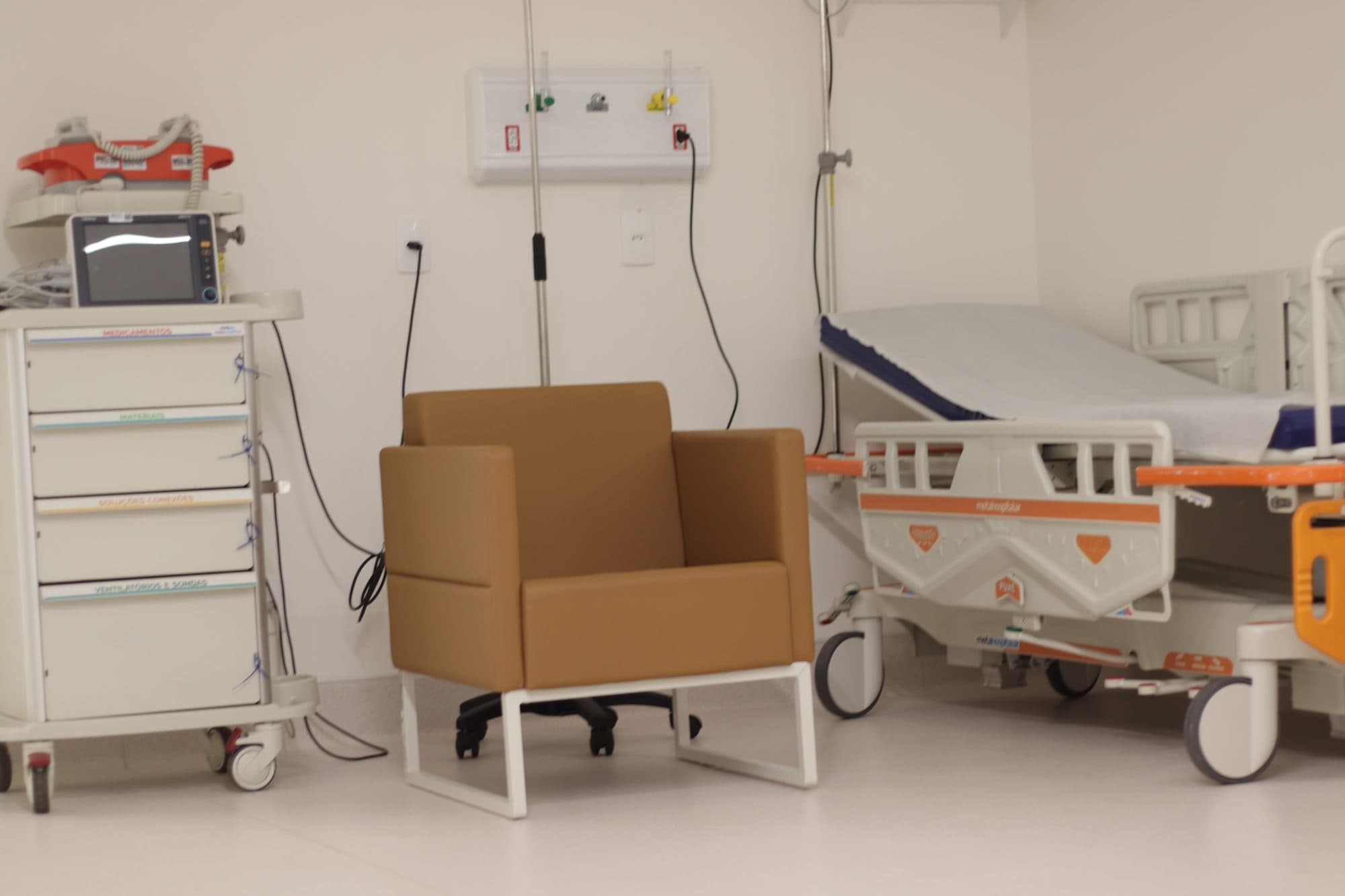 Sala de tratamento com poltrona, maca, carrinho de medicamentos e monitor multiparamétrico